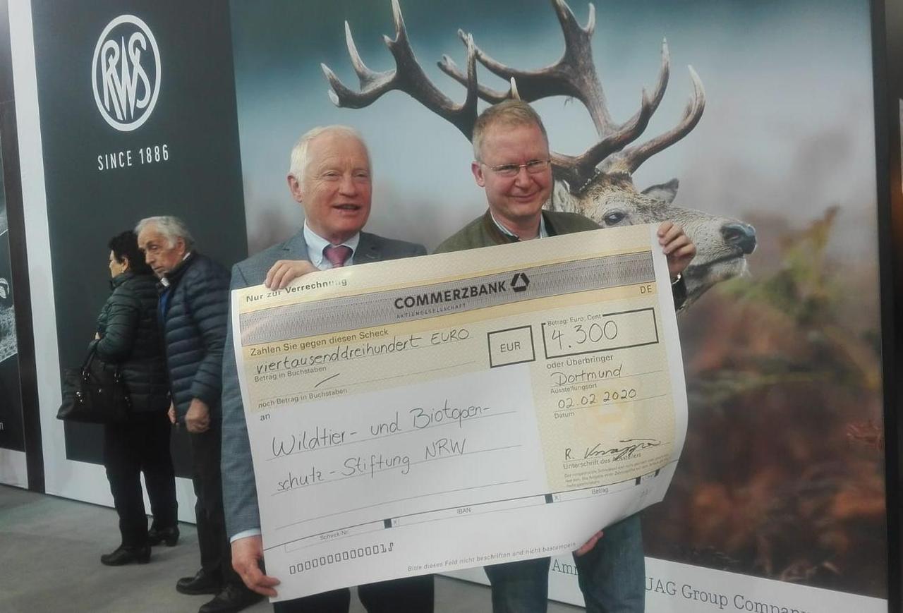 RWS spendet 4.300,- EUR für die Wildtier- & Biotopschutz-Stiftung NRW Scheckübergabe mit Papierscheck vor RWS Wand auf der Jagdmesse "JAGD & HUND"
