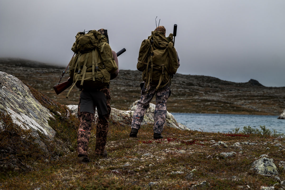 Zwei Jäger in Tarnkleidung laufen mit Rucksäcken durch felsige Landschaft 